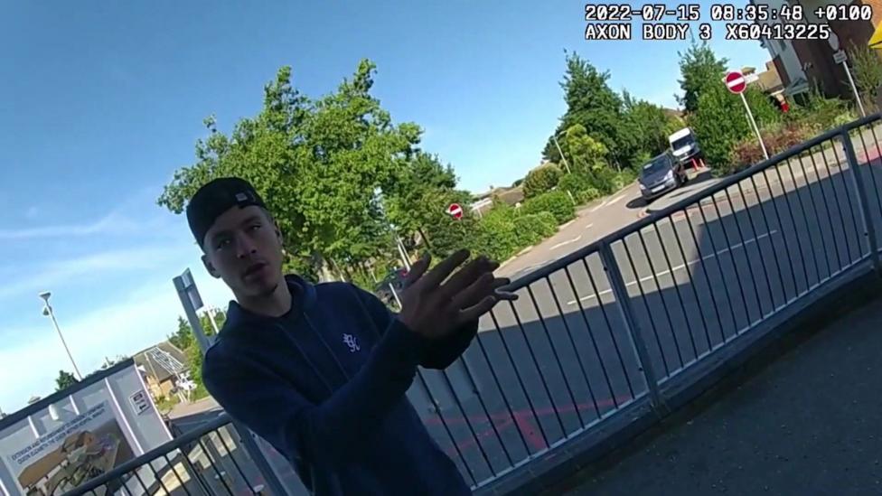 Tyla Wharmby wearing a dark hooded top and a backwards facing baseball cap gesticulates as he is recorded on a police bodyworn camera. A date in the corner of the image reads 15 July 2022.