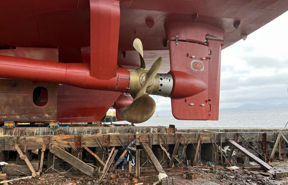 A large metal propeller on the underside of a red painted ship which is sitting on a slipway