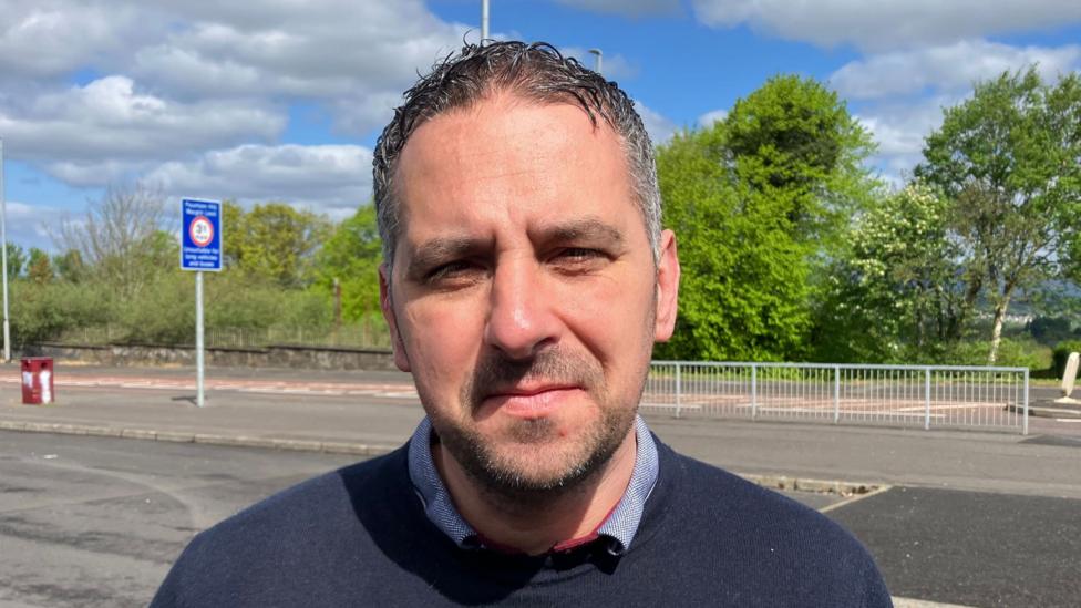 Sinn Féin councillor Christopher Jackson has short brown hair and is wearing a navy jumper and a blue shirt. He is standing in the middle of a street.