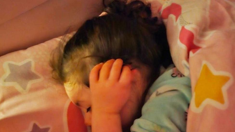Grace Miller in her bed, with her head resting on a pillow. She has her ears covered with her hands to try and block out any noise. 