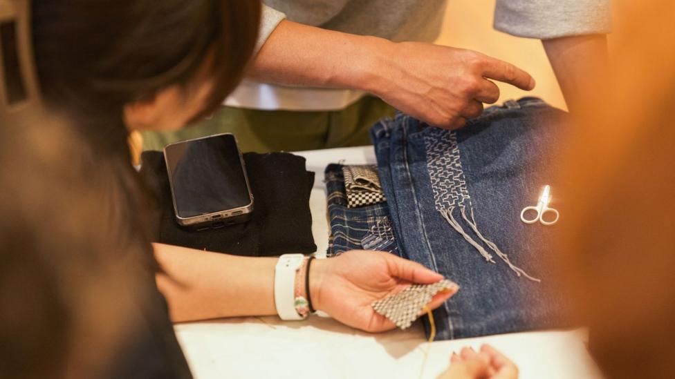 Close up of people collectively looking at some decorative white stitching on a pair of jeans. Only hands and out of focus sides of heads are visible.