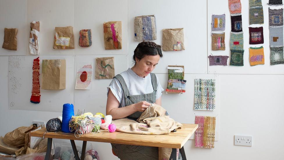 Woman mending at a table with samples on the wall behind her