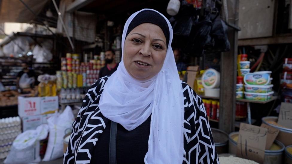 A woman in her 50s wears a white hijab and looks to the camera, in front of an outdoor market stand selling food items