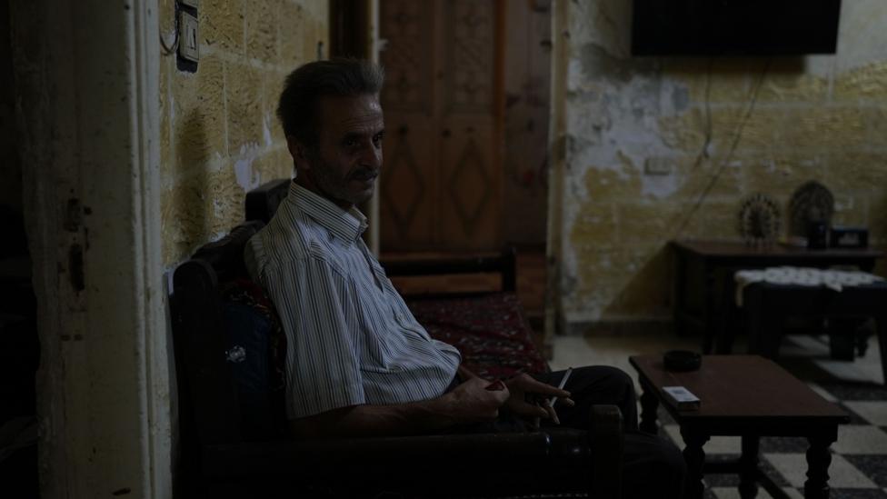 A man wearing a striped shirt sits in his living room in darkness