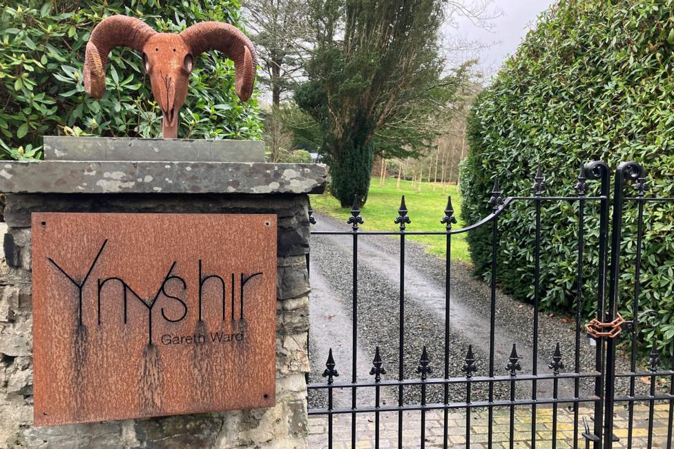 Front iron gate and aged iron sign saying Ynyshir with driveway beyond and hedges