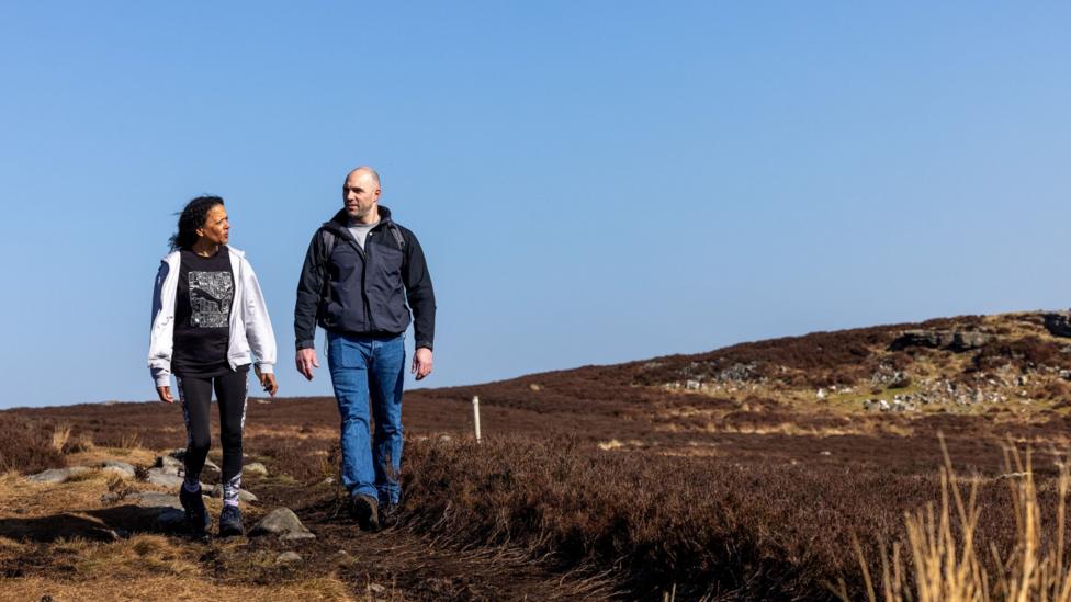 A woman and a man walk along a trail on the Simonside Hills. The woman, on the left, has dark hair and is wearing a white jacket with a black top and pants. Her companion has a shaved heard and is wearing a grey waterproof jacket and blue jeans.