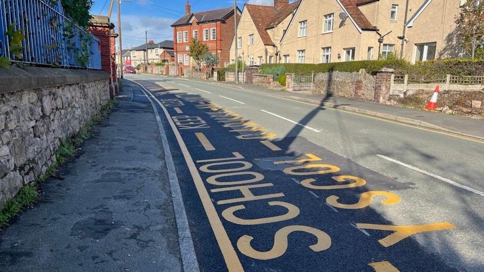 A road with houses on one side and a path on the other way with a wall and a blue, rusted fence. There is yellow painted signage on the road showing a school is there.