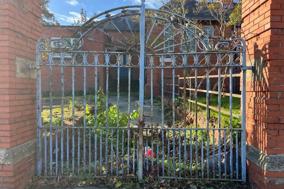 Blue rusted gates closed outside a red brick building. There is overgrown shrubbery behind.
