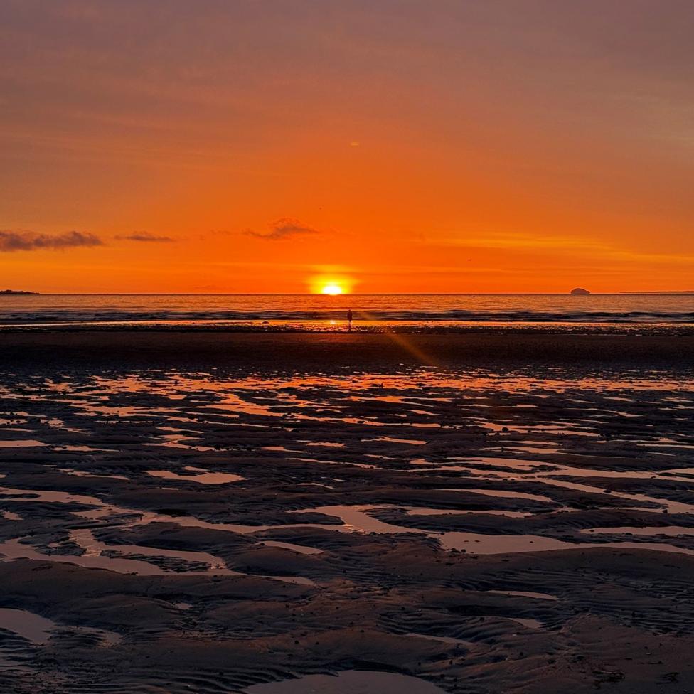 A solitary figure on a beach at sunrise with an orange glow on the distant horizon.