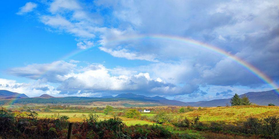 A rainbow arching over some sunlit countryside under a cloudy blue sky with hills in the distance.