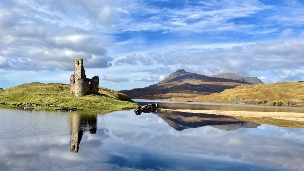 Ruins of a castle reflected in a loch, with a hillside in the background.