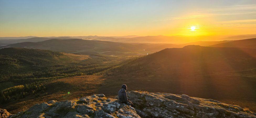 A young top sitting at a hillside summit watching an orange sunset.