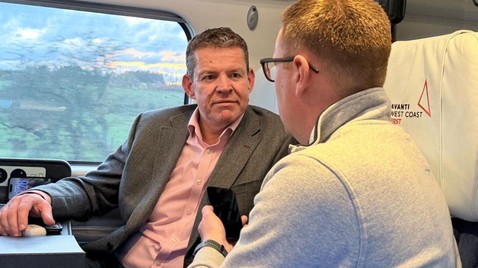The image shows Rhun ap Iorwerth speaking to a journalist on a train. He is wearing a suit and an open collar pink shirt.