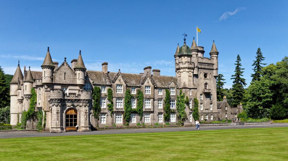 Large grey stone castle with turrets and towers, partially covered in green climbing ivy, standing behind a wide manicured lawn under a clear blue sky. A flag flies from the central tower, with a few visitors visible near the entrance.