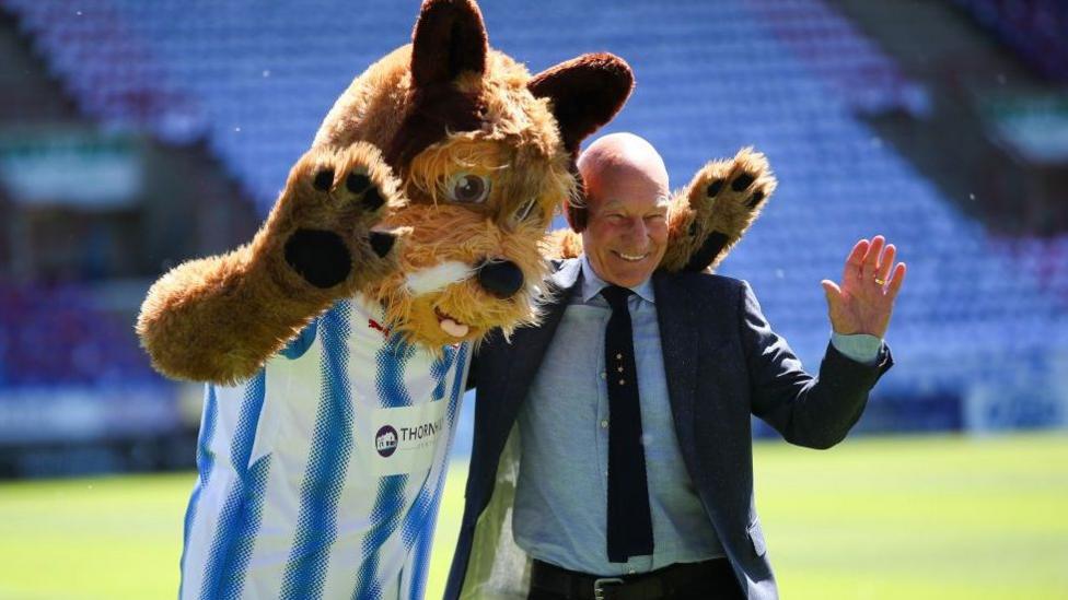 Patrick Stewart - smiling man wearing a suit stood with a dog mascot dressing in a blue and white football top. 