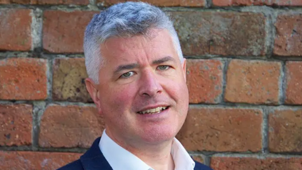 Man with short grey hair wearing a white shirt and dark jacket, standing in front of a red brick wall and smiling slightly at the camera.