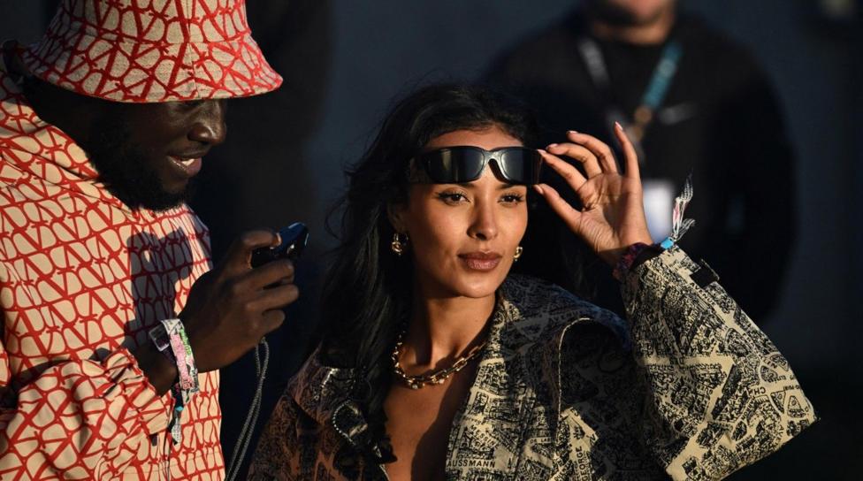 Maya Jama holding sunglasses above her eyes and standing next to rapper Stormzy. Maya wears a cream and black jacket with map prints all over it. Stormzy wears a read and cream suit with a bucket hat and he is holding a camera.