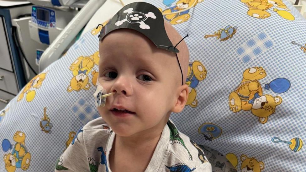 A young boy wearing a pirate hat. he has a feeding tube on his nose and is sat in a hospital bed with cartoon patterned bedding behind him.