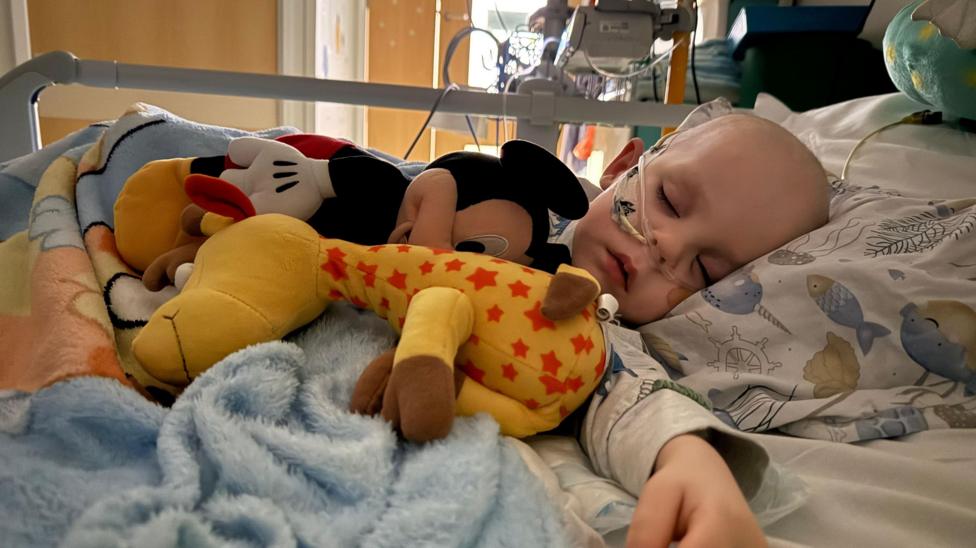 A little boy sleeping in a hospital bed with a soft toys by his side.
