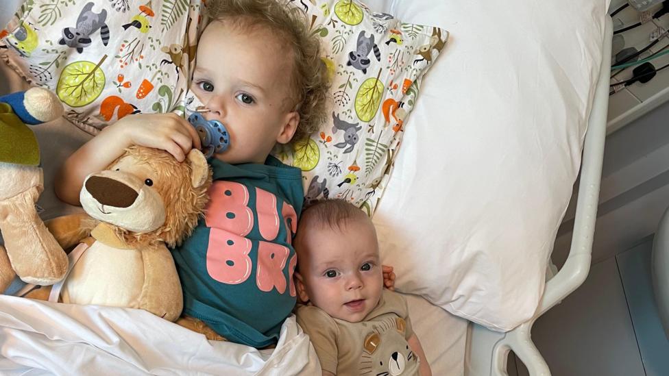 A young boy and a baby boy laying in a hospital bed with stuffed toys.