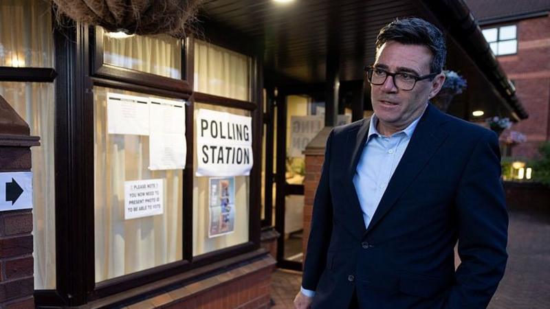 Andy Burnham in a dark suit and open necked blue shirt, walks past a sign saying Polling Station