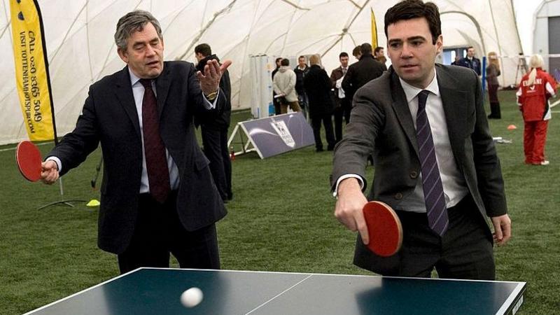 Gordon Brown and Andy Burnham making up doubles pair at a table tennis table in a tent at a sports ground in 2009. They are both wearing suits.