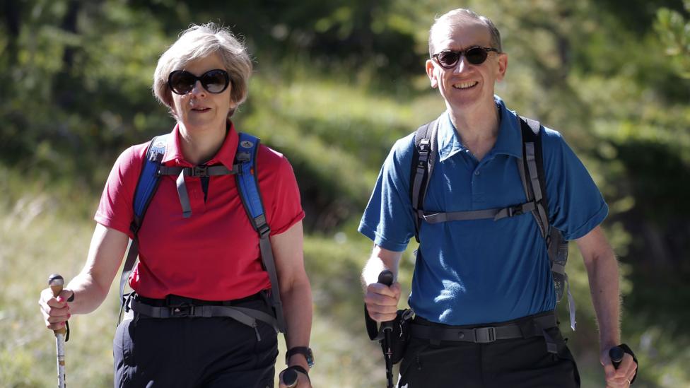 Theresa May wearing outdoor gear and sunglasses strides along a country road in the Swiss Alps with husband. Both are holding hiking polls.