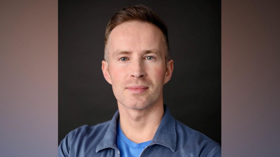 A man with short brown hair wears a blue t-shirt and and navy jacket as he looks to the camera. 