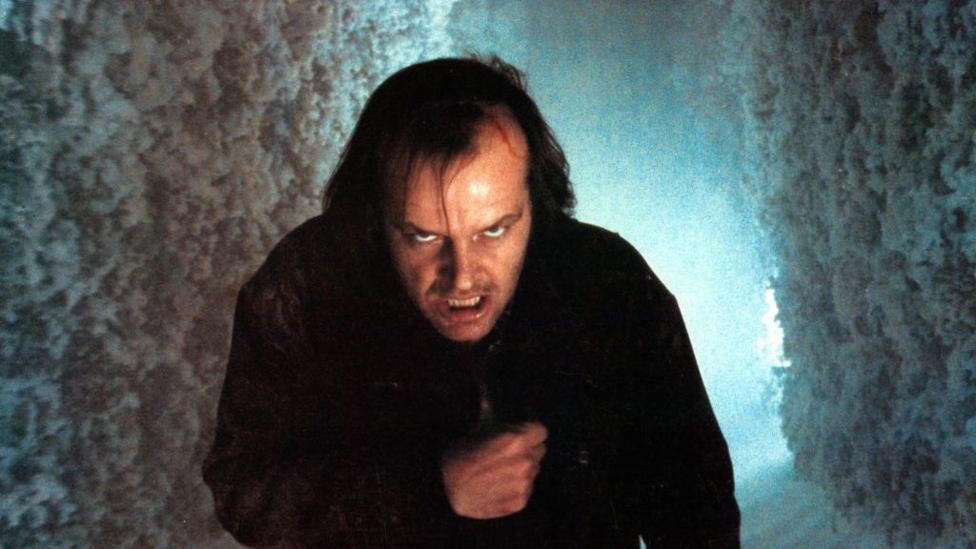 A still of a man with medium length brown hair walking through a snow covered maze. He wears a dark jacket and has a cut on his head. 