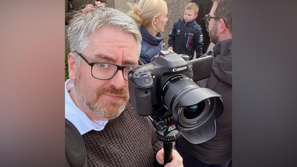 A man wears a brown jumper over a white shirt. He takes a selfie whilst holding a black Canon camera. He wears black glasses, with clear lenses and has short grey hair. 