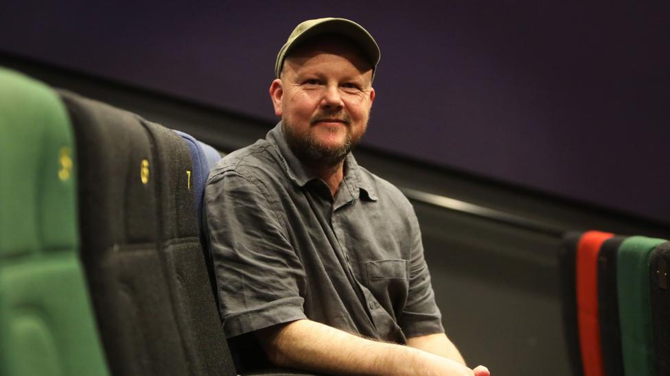 A man sits on a blue chair facing the camera. He wears a green cap and black short sleeve shirt. He is sitting in a movie theatre with purple walls and red, green, blue and black chairs. 