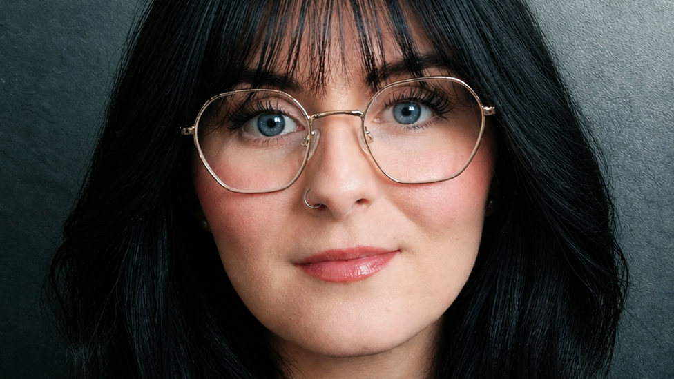 A woman with dark hair and a fringe. She has striking blue eyes and is wearing glasses.
