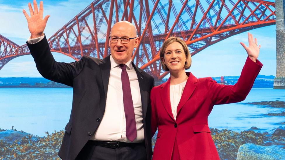 John Swinney and Jenny Gilruth standing in front of a background with the Forth Rail Bridge on it.