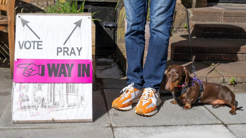 A sign outside a polling station in Scotland. The lower half of a person wearing blue jeans and orange training shoes is to its left, alongside a small brown sausage dog.
