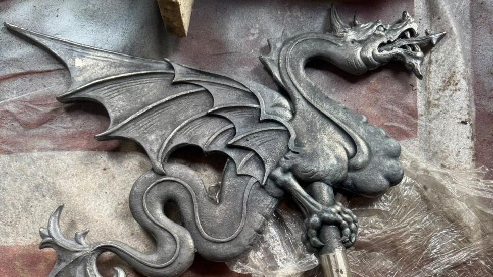 A decorative iron dragon on a pole. It is side on with large wings, a tail, and it's tongue out.