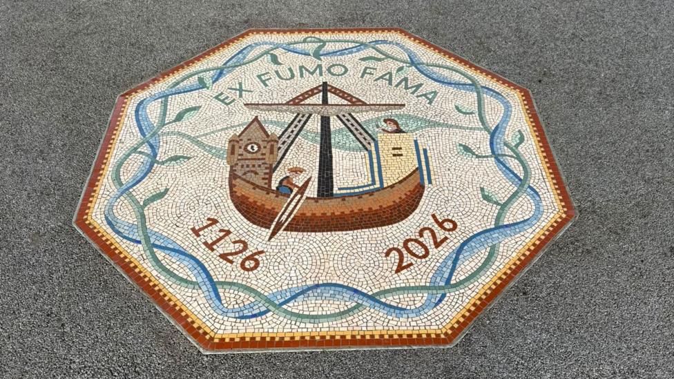 The mosaic is octagonal with a red and yellow border and then another interlocking blue and green border. The green border also has leaves. The words 'EX FUMO FAMA' are at the top in capitals and at the bottom is '1126' and '2026'. In the middle is a boat with buildings on it.